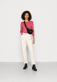 Bolsa crossbody preta com fechos, usada sobre uma camisa de manga longa rosa e combinada com calças brancas de cintura alta e botas de tornozelo pretas.