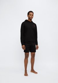 Μαύρο φούτερ με κουκούλα και κόκκινο λογότυπο καρδιάς στο στήθος, σε συνδυασμό με μαύρο σορτς. Και τα δύο κατασκευασμένα από μαλακό ύφασμα, με άνετη εφαρμογή και μίνιμαλ σχεδιασμό.