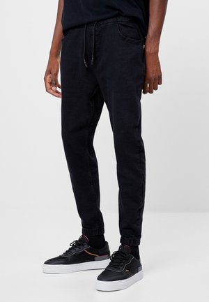 Uomo con pantaloni jogger neri con risvolti elastici e sneaker nere con suola bianca, in piedi su uno sfondo semplice grigio chiaro.