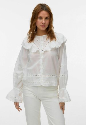 Kobieta ubrana w białą bluzkę z dużym falbanowym kołnierzem i mankietami, ozdobioną ażurowymi detalami, oraz białe spodnie, stojąca na jednolitym tle.