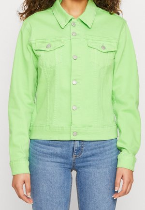 Femme portant une veste en jean verte vif à boutons avec poches poitrine et un jean bleu, vue du cou aux hanches, debout devant un fond blanc.