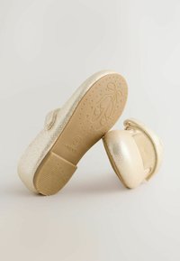 Goldene Ballerinas mit einer strukturierten, metallischen Oberfläche, transparentem Riemen und flacher Gummisohle mit Stern- und Ballerinaschuh-Muster.