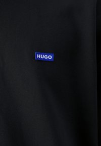 Tissu noir avec une petite pièce rectangulaire bleue comportant le texte blanc "HUGO" cousu près du centre.