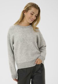 Ung kvinde smilende, iført en lys grå sweater med chevronmønster og sorte læderbukser, med den ene hånd i lommen foran en hvid baggrund.