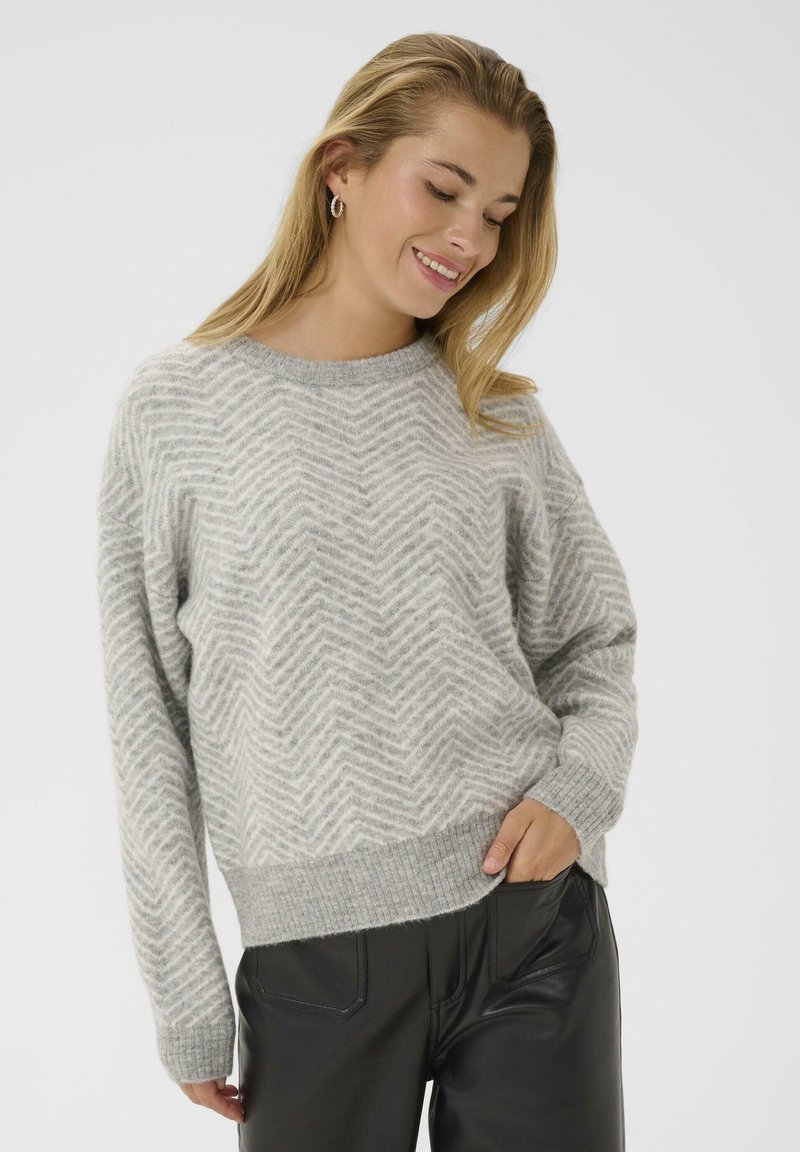 Ung kvinde smilende, iført en lys grå sweater med chevronmønster og sorte læderbukser, med den ene hånd i lommen foran en hvid baggrund.