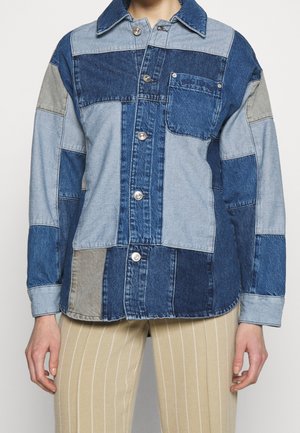 Kvinde iført en patchwork denimjakke med forskellige nuancer af blå og grå, sølvknapper og beige bukser med tynde striber.