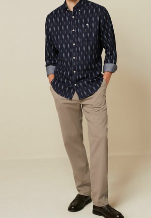 Chemise bleu marine à boutons avec un motif géométrique, manches retroussées, pantalons khaki clair et mocassins noirs. Textures subtiles et détails contrastés.