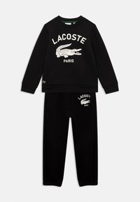 Sæt bestående af sort sweatshirt og bukser med hvid Lacoste-logo og krokodillegrafik på brystet og venstre lår, designet til afslappet brug.