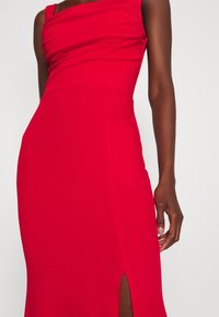 Robe rouge avec un col drapé et un corsage ajusté. Elle présente une fente haute sur la cuisse d'un côté, réalisée dans un tissu lisse et extensible.