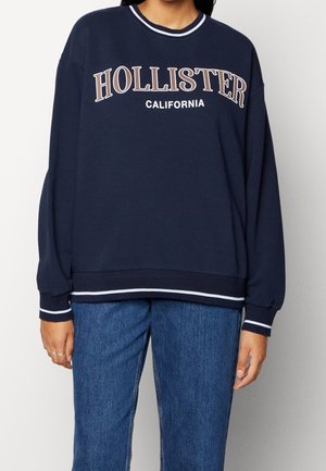 Person iført en navyblå Hollister California sweatshirt med hvide og grå bogstaver, kombineret med blå jeans, stående mod en ensfarvet baggrund.