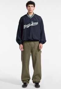 Jaunas vyras vilki tamsiai mėlyną "Guess Jeans" megztinį su baltomis ir žaliomis juostelėmis bei laisvas alyvuogių žalios spalvos krovinių kelnes ir juodus sportinius batus.