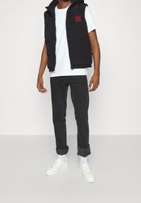 Schwarze Steppweste mit Kapuze und rotem "HUGO"-Logo, getragen über einem weißen T-Shirt, kombiniert mit schwarzen Jeans und weißen Sneakers.