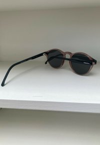 Gafas de sol redondas con un marco rosa translúcido, lentes oscuros y patillas negras. Incluye un logotipo, marcado CE y etiqueta de protección UV Cat 3.
