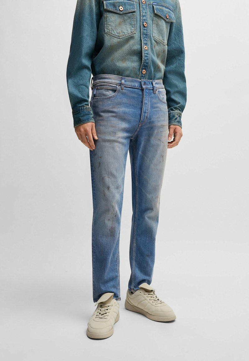Light blue denim jeans with a faded finish, featuring subtle stains, classic five-pocket design, and straight leg shape. Paired with off-white sneakers.
