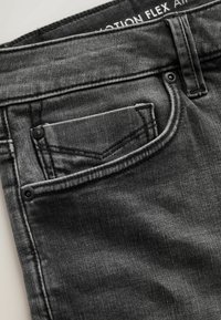 Gros plan sur un jean en denim noir montrant la poche avant, la petite poche à monnaie avec rivets, et la ceinture avec coutures visibles et texture du tissu.