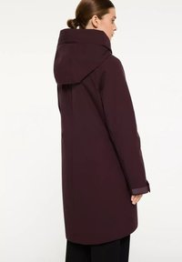 Vrouw in een lange, donkerpaarse jas met een grote capuchon, staand in zijaanzicht tegen een effen witte achtergrond.