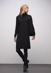Robe longue noire à manches longues avec un col haut et une coupe décontractée, associée à des bottines noires. Texture lisse avec une légère élasticité.