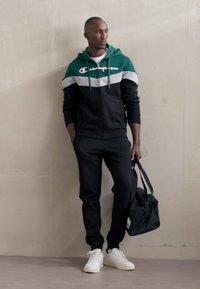 Chándal negro con una chaqueta con capucha verde y gris, que presenta un cierre de cremallera en la parte delantera y un logo. Zapatillas blancas y una bolsa negra complementan el conjunto.