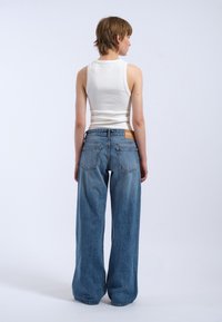 Jean large bleus avec une décoloration, cinq poches et une étiquette en cuir marron sur la taille au dos. Assortis à un débardeur blanc.