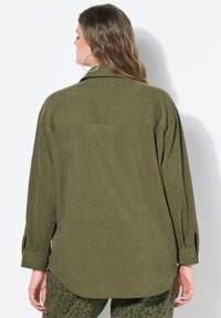 Olivenfarvet oversized skjorte med lange ærmer, rygpanel og knaplukning ved håndleddene. Blød tekstur, buet kant, parret med mønstrede underdele.
