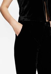 Schwarzes Samtoberteil mit einem kurzen Design und vertikalen Taschen, kombiniert mit passenden schwarzen Samthosen mit einer glatten Textur.