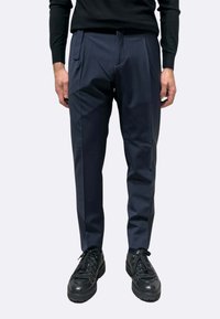 Pantaloni a sigaretta blu navy in tessuto liscio con tasche laterali, abbinati a sneaker nere con lacci texture e finitura lucida.