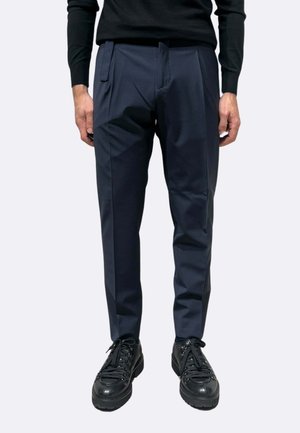 Pantaloni a sigaretta blu navy in tessuto liscio con tasche laterali, abbinati a sneaker nere con lacci texture e finitura lucida.