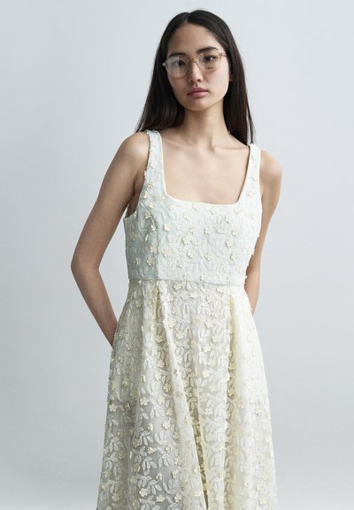 Robe blanche brodée de fleurs avec un décolleté carré et un design en couches. Présente des fleurs délicates sur un filet transparent et un corsage structuré.