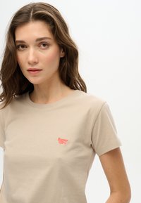 T-shirt en coton beige à manches courtes, arborant un petit logo rouge brodé "Super Dry" sur le côté gauche de la poitrine.