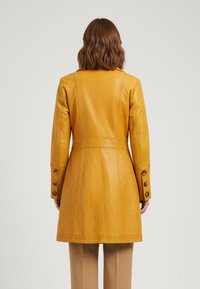 Mustard yellow leather coat with a tailored fit, prominent seam detailing, and buttoned cuffs. Smooth texture and knee-length design.
