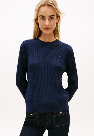 Blonde vrouw met een gestructureerde marineblauwe trui met lange mouwen en een donkerdenim, staand met handen achter haar rug, neutrale achtergrond.