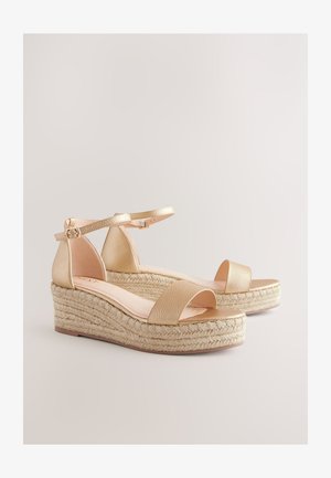 Złote sandały na klinie z paskiem wokół kostki, z fakturowaną cholewką i plecioną platformą z juty. Otwarty przód i gładka wkładka.