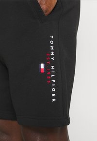 Pantalones cortos negros con una textura suave. Presenta el bordado "TOMMY HILFIGER" en blanco y "EST. 1985" en rojo, junto a un pequeño emblema de bandera.