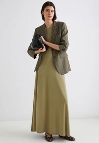 Un maxi dress verde oliva abbinato a un blazer verde oliva su misura con bottoni. La donna tiene una borsa nera e indossa scarpe con stampa leopardata.
