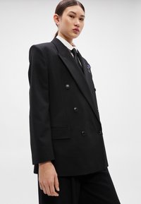 Μαύρο διπλόμπιστο σακάκι με γιακάδες με χαραγμένη εγκοπή, εμπρόσθια κουμπιά και πλαϊνές τσέπες. Φορεμένο πάνω από λευκό πουκάμισο και μαύρη γραβάτα, με διακοσμητικό καρφίτσα.