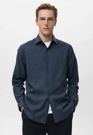 Camisa de botones azul oscuro con una tela texturizada, mangas largas y cuello puntiagudo, combinada con una camiseta blanca por debajo.