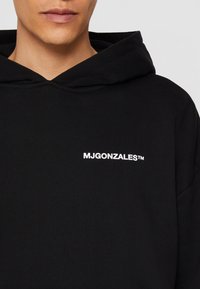 Homme portant un sweat à capuche noir avec le texte blanc "MJGONZALES™" centré sur la poitrine, lèvres et partie inférieure du visage visibles.