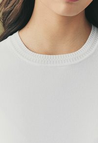 Pull en tricot blanc avec un col rond texturé et côtelé. Tissu doux avec un aspect lisse et léger. Aucun motif ni matériel visible.