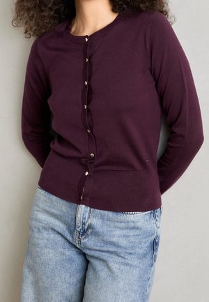 Kvinde iført en tætsiddende mørklilla cardigan med guldknapper og lyseblå højtaljede jeans, stående mod en ensfarvet grå baggrund.
