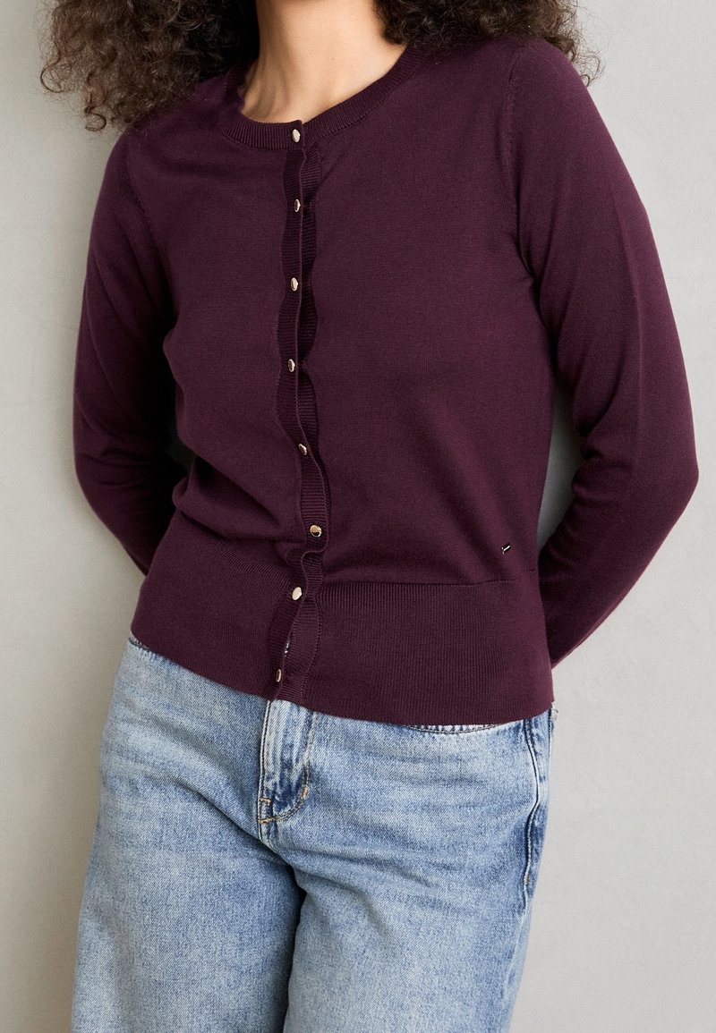 Femme portant un cardigan ajusté violet foncé avec des boutons dorés et un jean taille haute bleu clair, debout devant un fond gris uni.