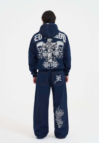 Granatowy dżinsowy hoodie i spodnie z grafikami czaszki, róży i krzyża. Wyróżniające szczegóły to kontrastowe białe szwy i nadruk smoka.