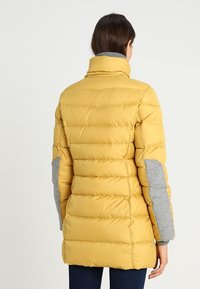 Gelbe Puffjacke mit hohem Kragen, horizontaler Steppung und grauen strukturierten Ärmeln. Das Design zeichnet sich durch einen mittellangen Schnitt aus.