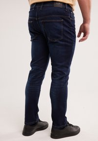 Mężczyzna w ciemnoniebieskich wąskich dżinsach i czarnych trampkach, stojący tyłem do kamery na jednolitym jasnym tle.