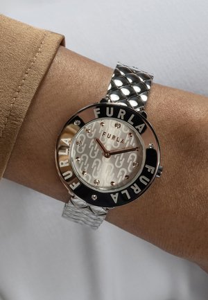 Srebrni sat od nehrđajućeg čelika sa teksturiranom narukvicom. Okrogli brojčanik sadrži svijetlu teksturu i crno slovo "FURLA", ukrašen kristalima.
