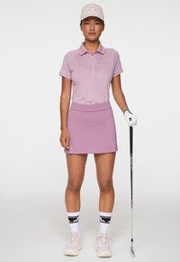 Giovane donna in abbigliamento da golf rosa che tiene una mazza da golf, indossando un cappellino, guanto, calzini e scarpe da ginnastica, in piedi davanti a uno sfondo neutro.