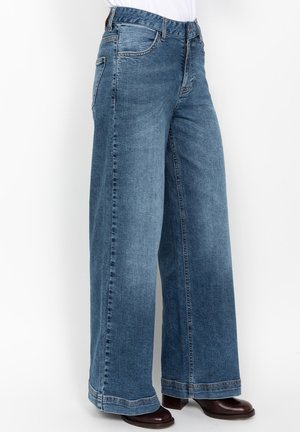 Jean en denim bleu à jambes larges porté avec des chaussures à talons marron foncé sur une personne, sur fond blanc.