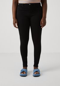 Persona vista con pantalones ajustados negros, una camiseta de rayas y zapatillas abiertas de dedo en color azul con cruces, de pie sobre un suelo blanco liso.