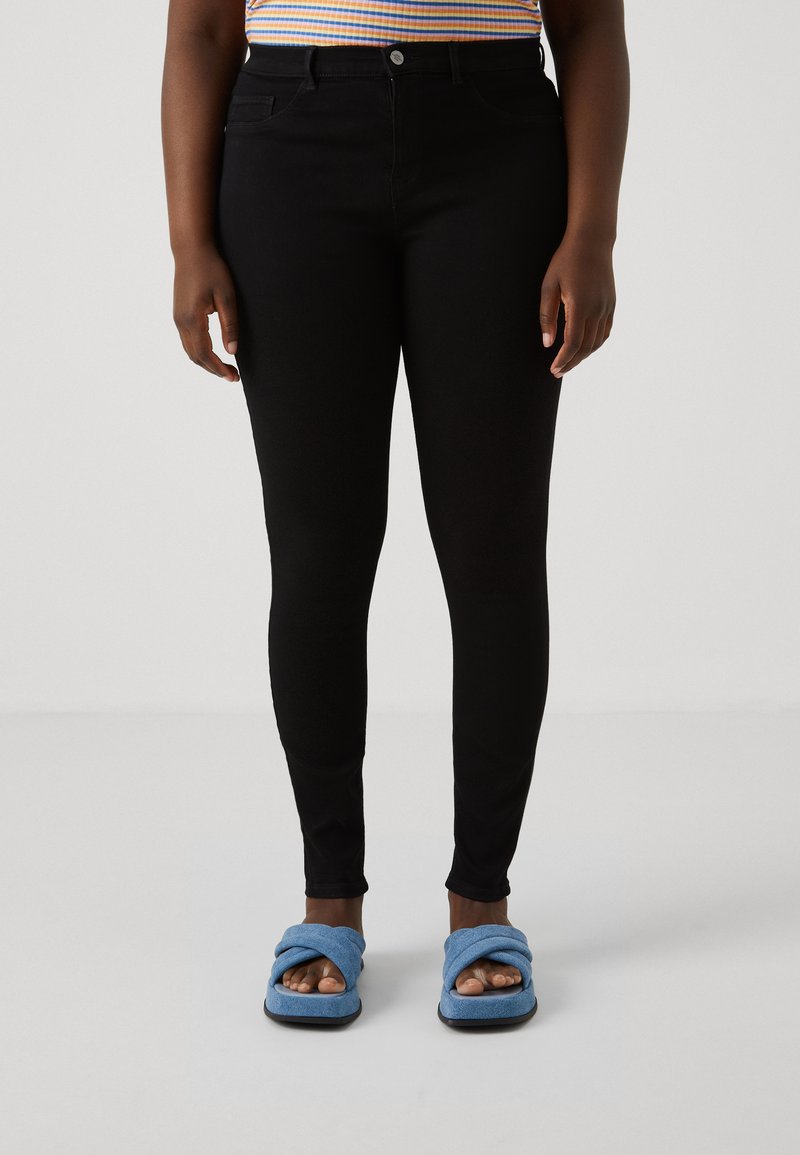 Persona vista con pantalones ajustados negros, una camiseta de rayas y zapatillas abiertas de dedo en color azul con cruces, de pie sobre un suelo blanco liso.