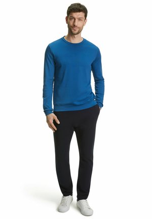 Blauw langemouwen shirt met een ronde hals, gecombineerd met donkere broeken en witte sneakers, gedragen door een staande man. Eenvoudige stof textuur.