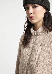 Beige fleecejack met een hoge kraag, volledige rits en een borstzak. Zachte textuur en minimalistisch ontwerp, met een donkere gebreide muts op het hoofd.
